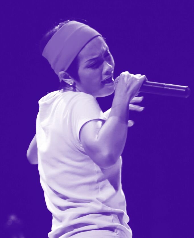 Person in a t-shirt holding a microphone, they appear to be singing or shouting into the mic. There is a purple shade to the entire photo.