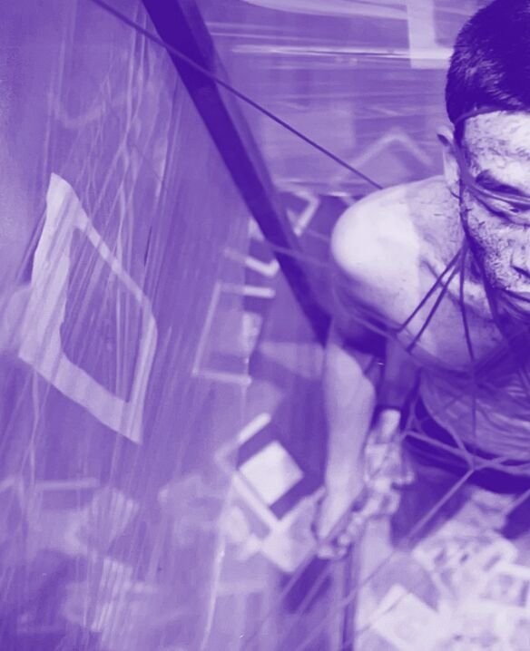The left half of a man's face is seen from above. He is wrapped in string. This string is connected to a large rectangular prism made of plastic that encapsulates the man. The photo has a purple hue 