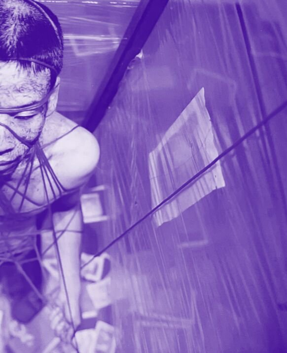 The right half of a man's face is seen from above. He is wrapped in string. This string is connected to a large rectangular prism made of plastic that encapsulates the man. The photo has a purple hue 