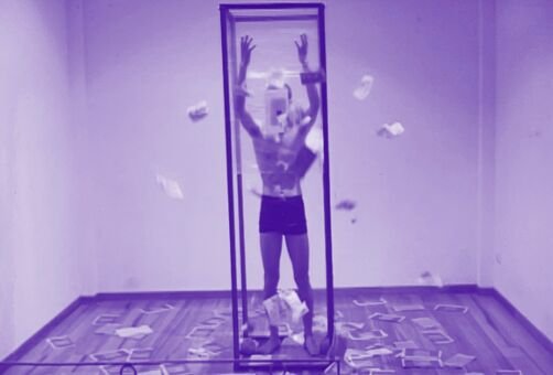 A man stand in a large vertical rectangular prism in his underwear. His arms are extended above his head. There is silicone wrapping around the prism. Papers fly all around him and settle on the floor. The image has a purple hue 