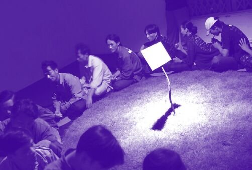 A group of people sit crossed legged on the floor facing each other's backs and form a circle. A tablet is elevated in the middle of the circle. The image has a purple hue.