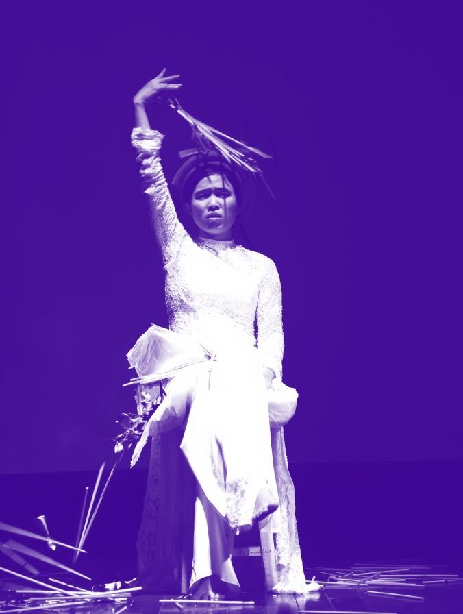 A woman in a white long sleeve dress holds what appears to be feathers above her head and allows them to fall onto her. The image has a purple hue