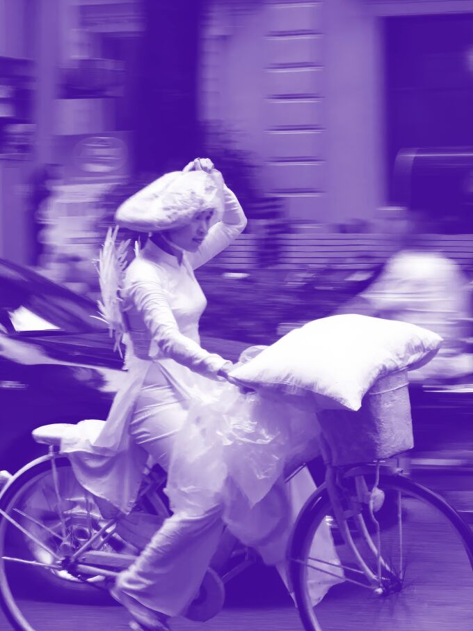 A woman in a white dress holds her white hat still as she rides a bicycle down the street. There is a pillow on top of the basket. There are feathers on her back. The image has a purple hue.