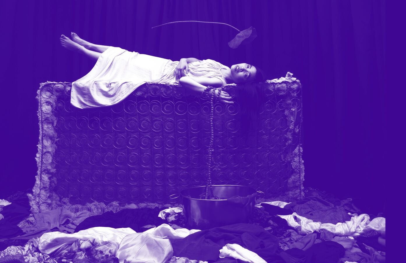 A woman in a white dress lies on top of an old mattress that is standing on its side. Clothes cover the floor below her. Beads are wrapped around her wrist and fall into a bucket on the floor. The image has a purple hue. 